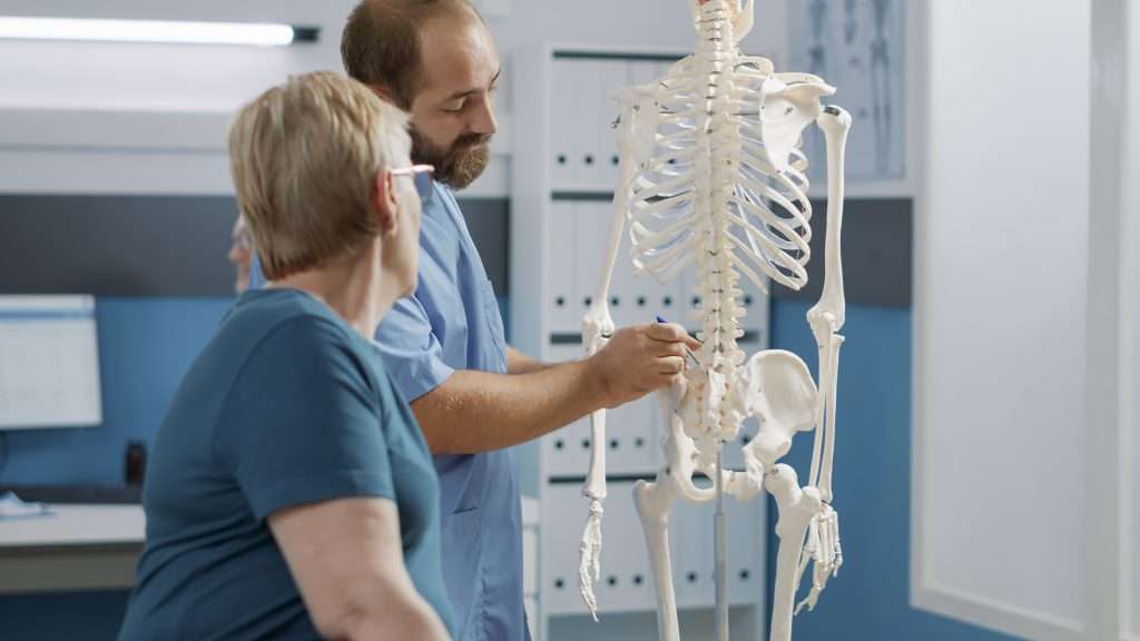 پزشک مرد با اشاره به اسکلت انسان برای نشان دادن نخاع و توضیح اختلالات مکانیکی در کابینه توانبخشی. متخصص در توضیح سیستم استخوان پشت برای فیزیوتراپی.
